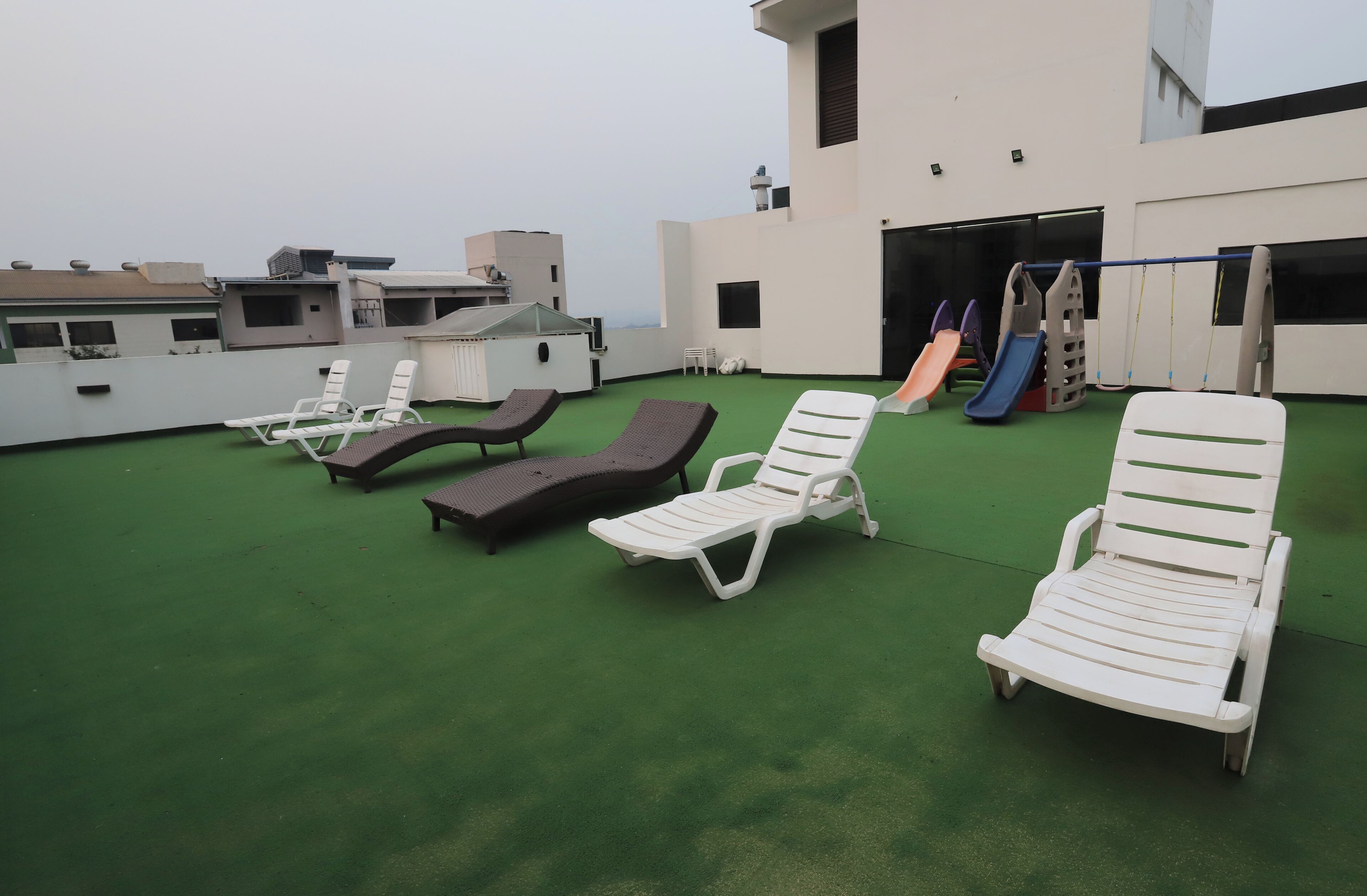 A rooftop with green artificial turf, white and brown lounge chairs, a children's slide, and a building with a flat roof.