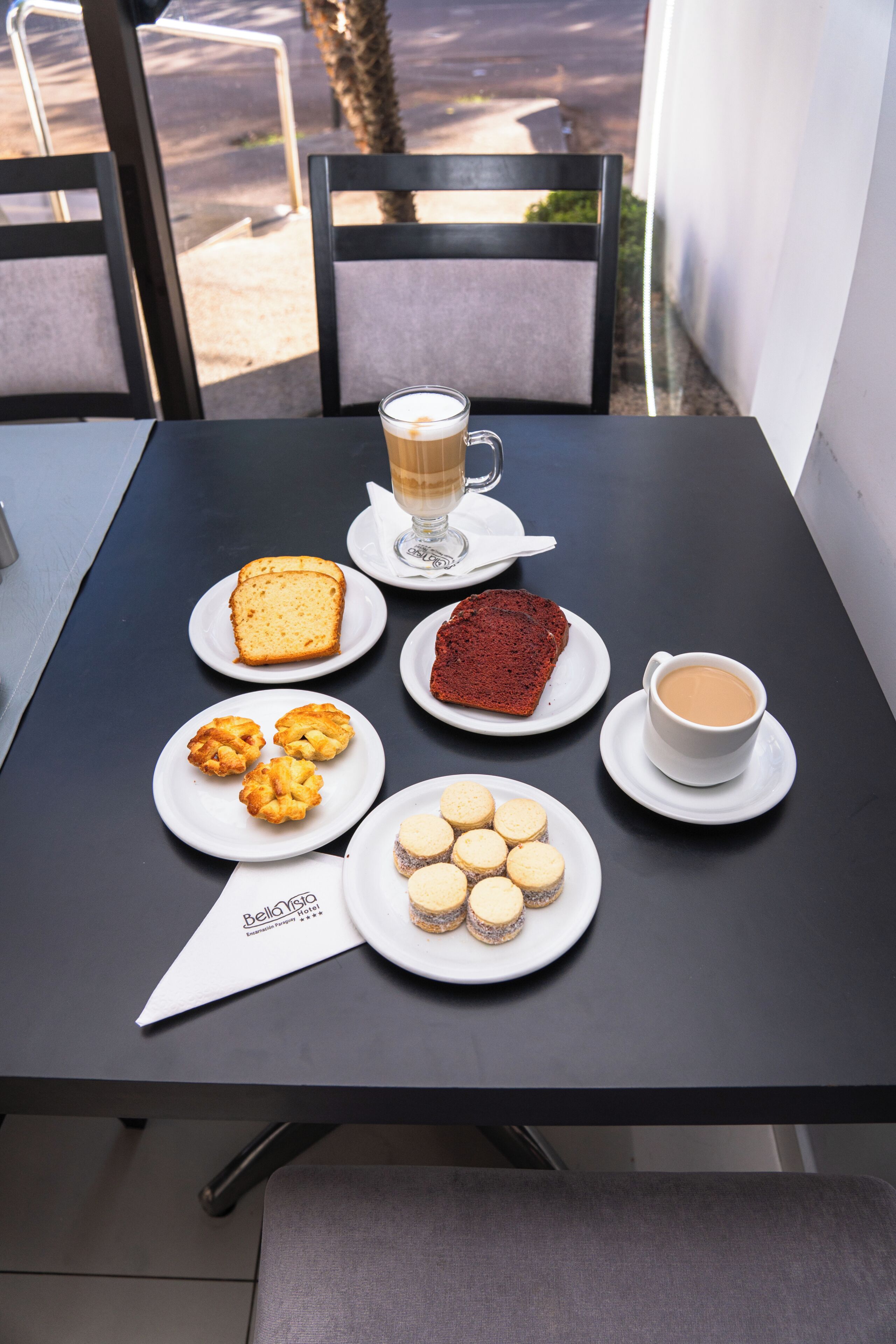 A table with various food items and a coffee cup, set against a window with a view of a road and trees.