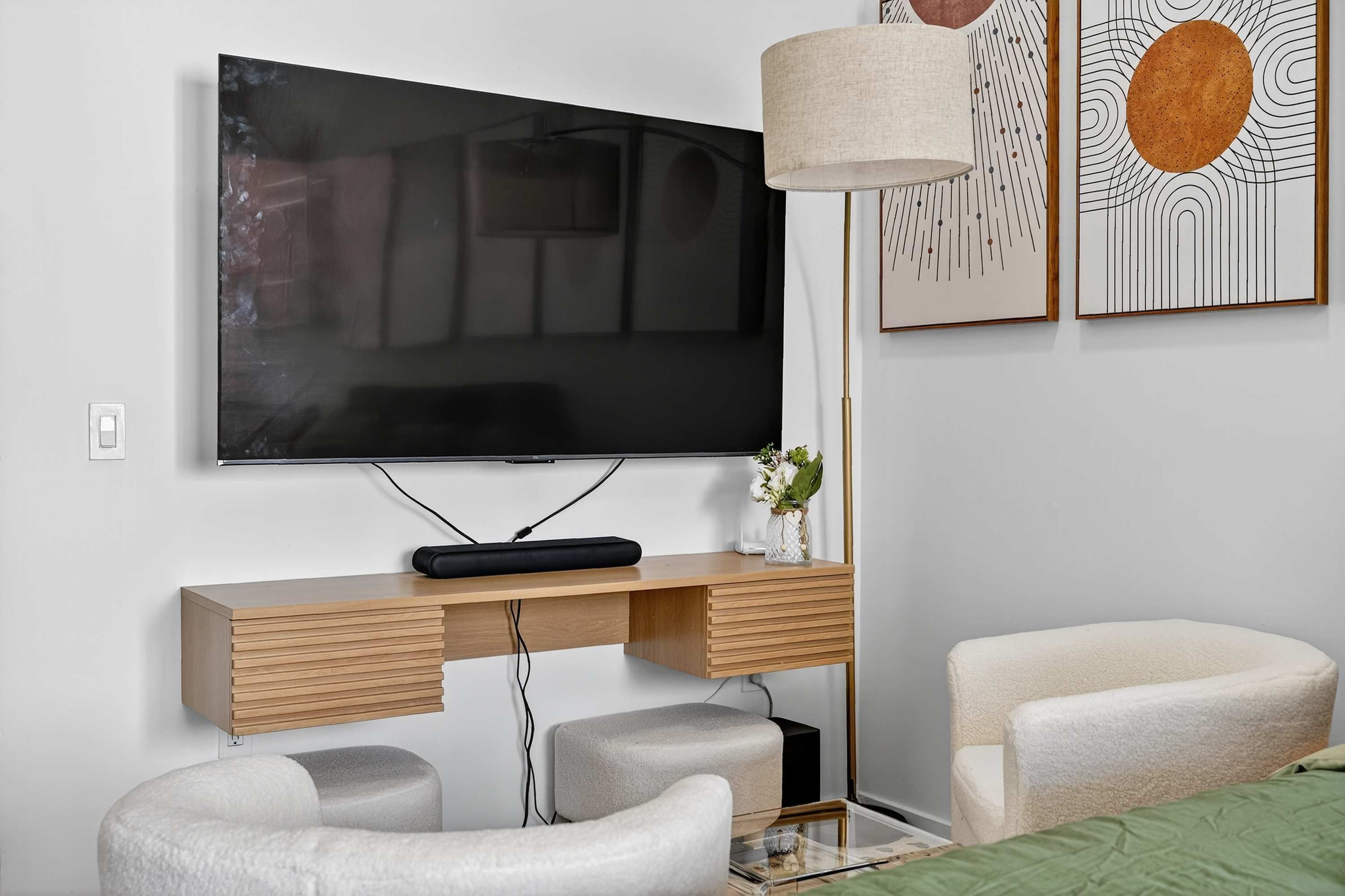 A modern living room with a flat-screen TV mounted on a wooden console, a white armchair, a glass coffee table, and a floor lamp.
