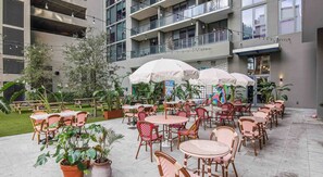 Ein möblierter Patio-Bereich mit Tischen und Stühlen, umgeben von Kübelpflanzen und einem Gebäude mit Balkonen.