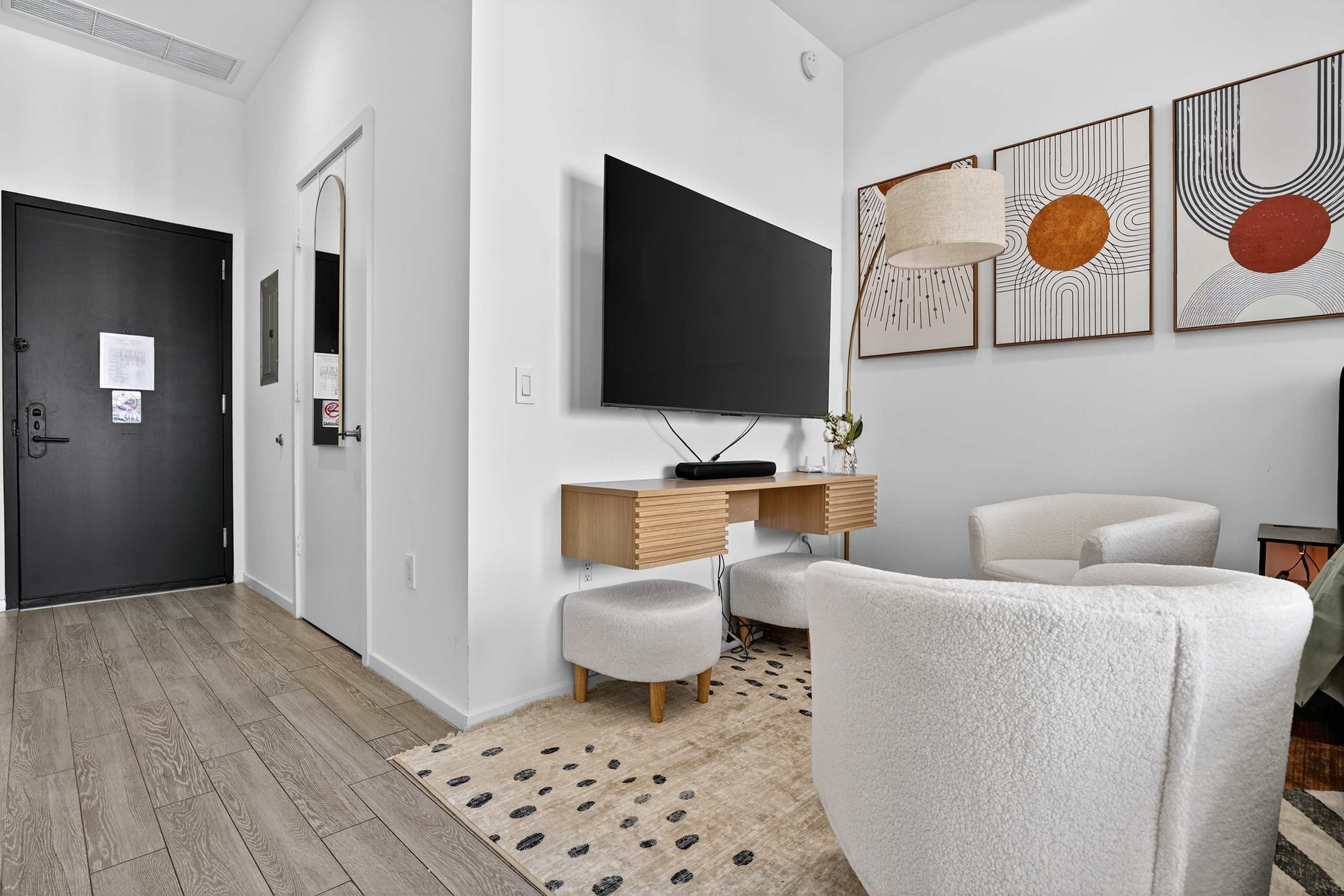 A modern living room with a flat-screen TV, a wooden desk, two white armchairs, and abstract wall art.