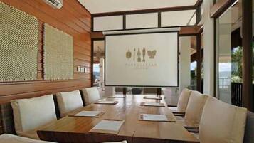 Uma sala de conferências com uma mesa comprida, cadeiras brancas e uma tela de projeção exibindo um logotipo.