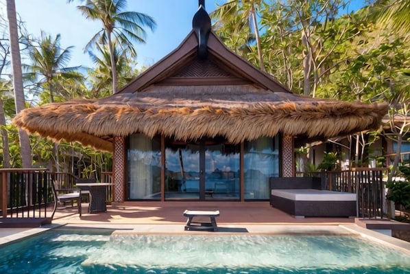 Estructura con techo de paja y piscina delantera, rodeada de palmeras.