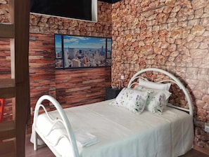 A bedroom with a stone wall, a bed with white bedding, and a cityscape painting on the wall.