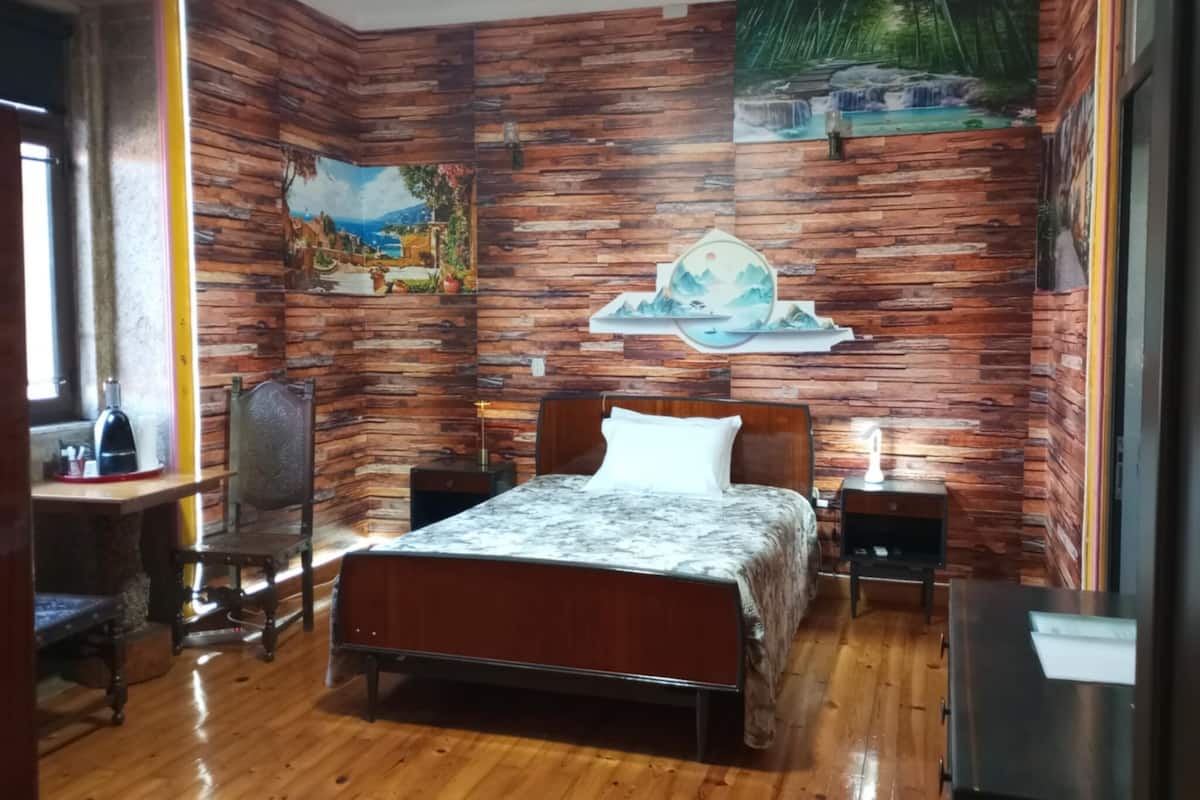 Ein Schlafzimmer mit Wandverkleidung aus Holz, einem Bett mit grau-weißem Muster, zwei Nachttischen mit Lampen, einem Sessel und einem Gemälde einer Wasserfall.'