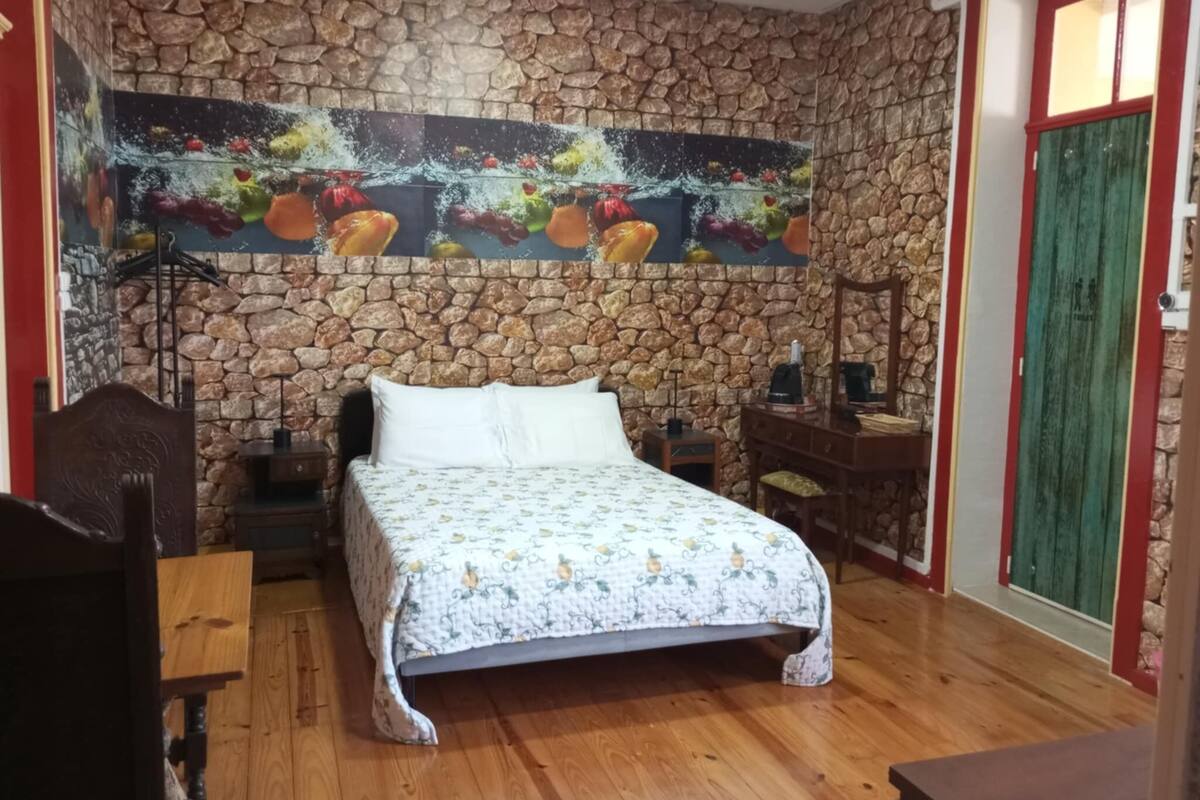Ein Schlafzimmer mit Steinsandsteinwand, einem Bett mit floralem Überwurf, einem Nachttisch, einer Kommode und einem Gemälde mit Früchten.