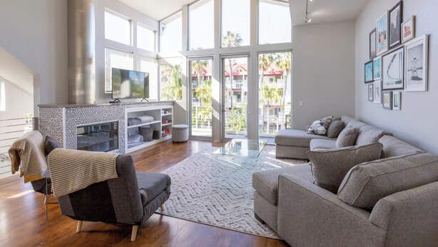 A spacious living room with a fireplace, a large TV, and a balcony with a view of palm trees and buildings. - Bayside Retreat - Bay Views in Walkable Coronado (Coronado)