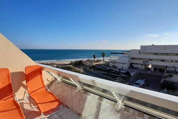 A balcony with orange chairs overlooking a beach and a parking area with buildings in the background.