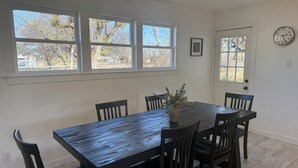 Una sala da pranzo con tavolo e sedie in legno scuro, ampie finestre con vista sugli alberi, una ventola a soffitto e un orologio appeso al muro.