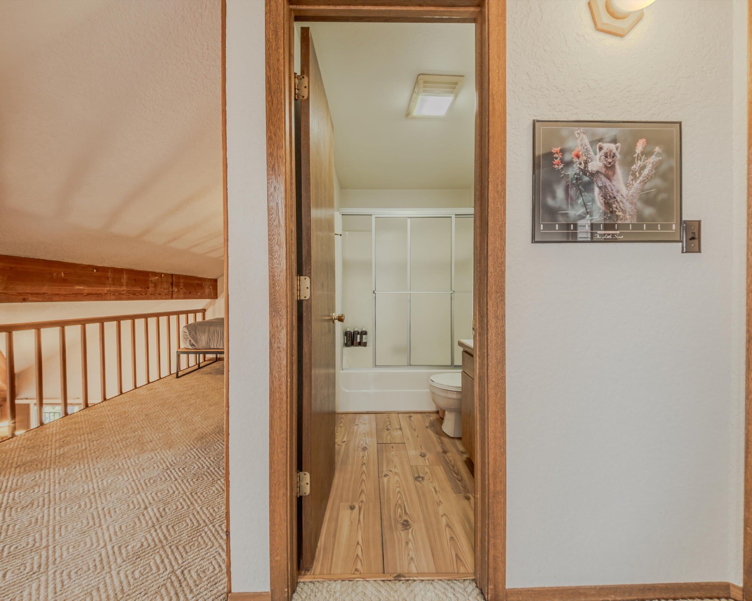 A hallway leading to a bathroom with a wooden floor, a white door, and a framed picture on the wall.