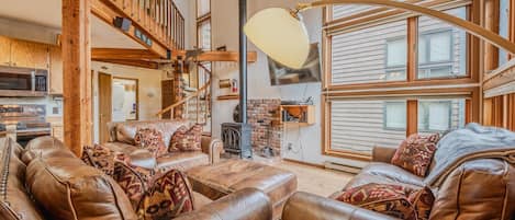 A spacious living room with leather sofas, a fireplace, a wooden staircase, and large windows.
