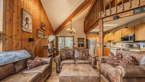A spacious living room with leather sofas and ottomans, wooden walls, and a kitchen area with wooden cabinets and modern appliances. - Sans Souci 1 3 Bedroom Ski In, Ski Out Retreat (Whitefish)