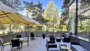 An outdoor patio with wicker furniture and yellow umbrellas, surrounded by trees and a building with large windows. - Hotel ILF (Prague)