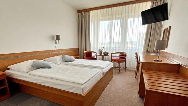 A hotel room with two beds, a desk, a chair, a TV, and a window with curtains. - Hotel ILF (Prague)