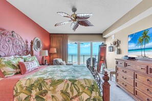 A bedroom with a bed, a ceiling fan, a dresser, and a large window with a view of the beach.