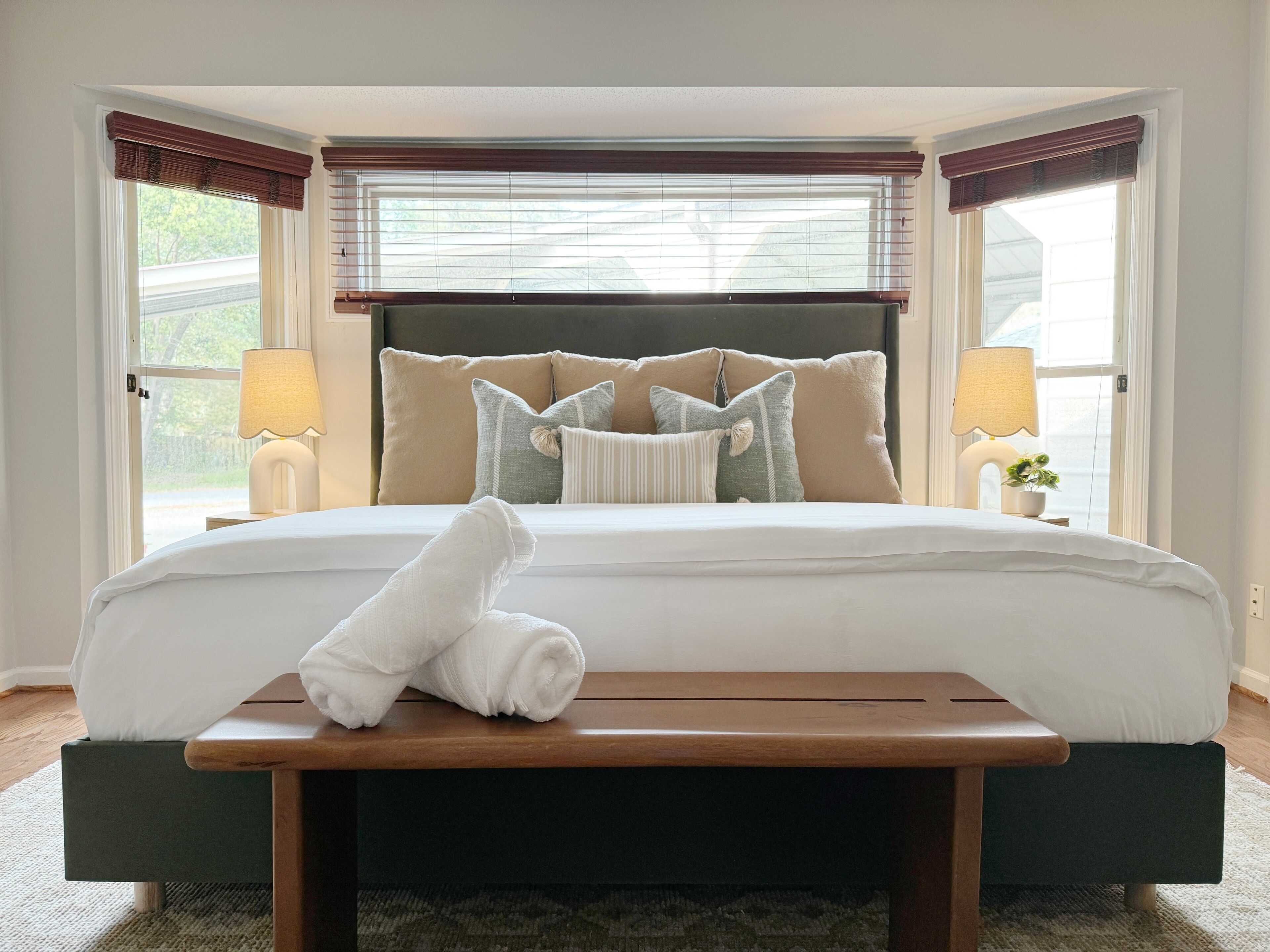 A neatly made bed with a rolled towel on a wooden bedside table, a headboard with multiple pillows, and two bedside lamps.