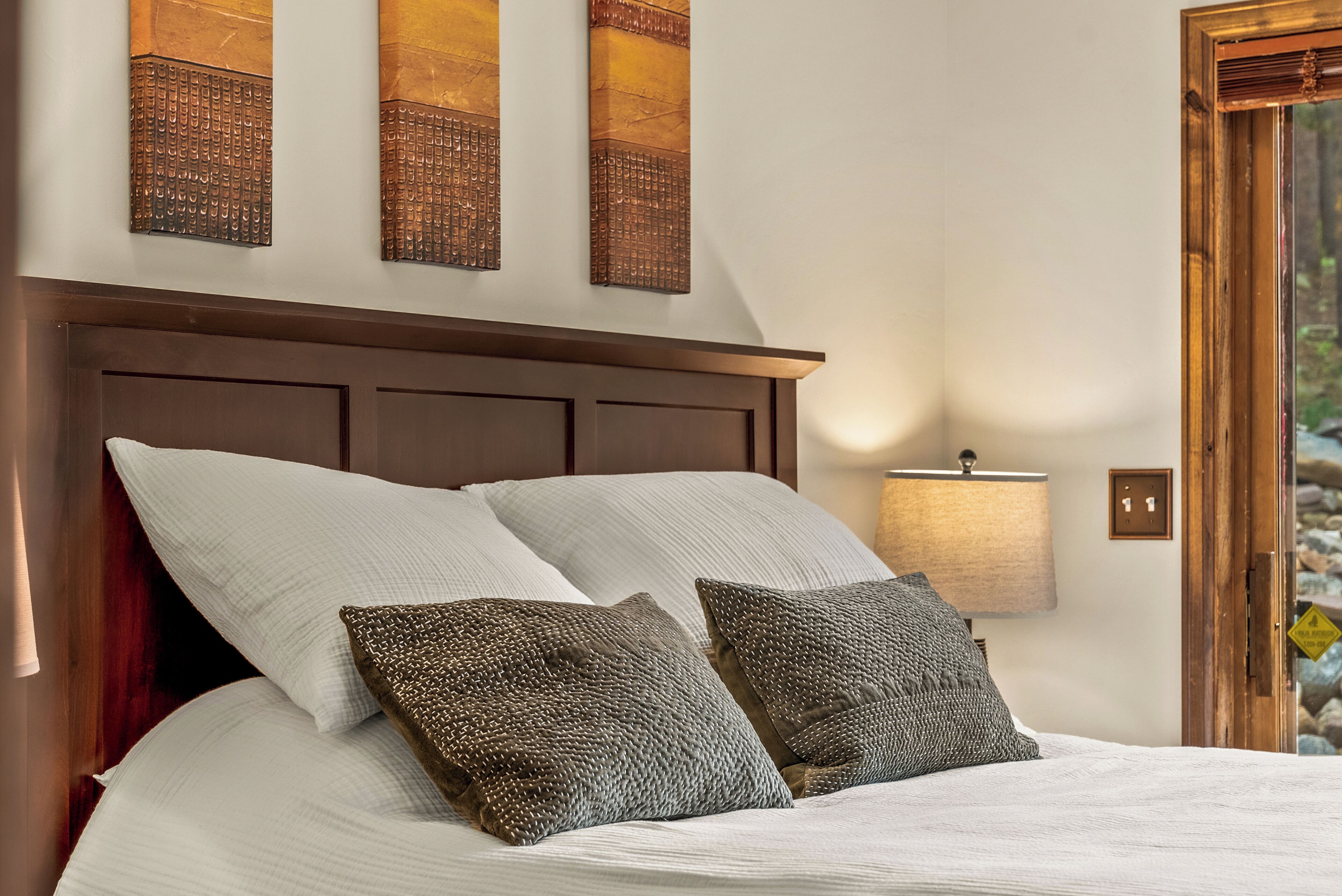 A neatly made bed with white linens and two patterned pillows, a wooden headboard, and a wall-mounted lamp.