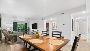 A dining area with a wooden table, chairs, and a large vase. There is a television on the wall and a window with curtains. - Cozy Creekside Condo with Resort-Style Amenities (Branson)