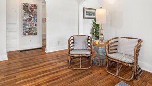 Una habitación con suelo de madera, dos sillas de mimbre con cojines, una lámpara y un cuadro en la pared.