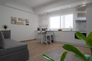 Eine moderne Küche mit weißen Schränken, ein Essbereich mit weißem Tisch und Stühlen sowie ein graues Sofa.