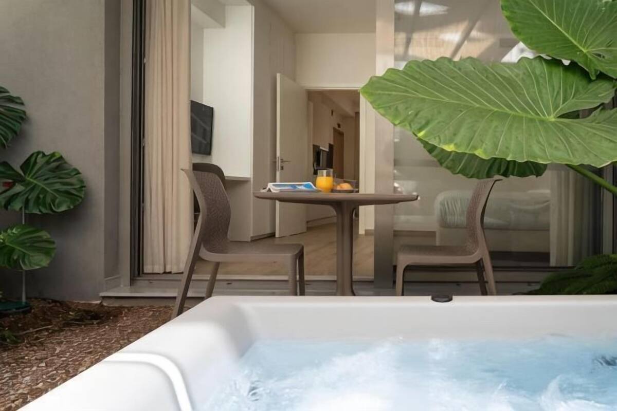 A hot tub with a table and chairs outside a building, surrounded by large plants.