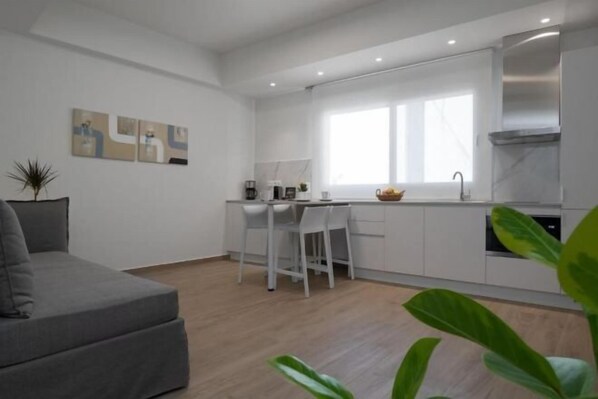 Una sala moderna con un sofá gris, un comedor con sillas blancas y una cocina con gabinetes blancos y un extractor de acero inoxidable.