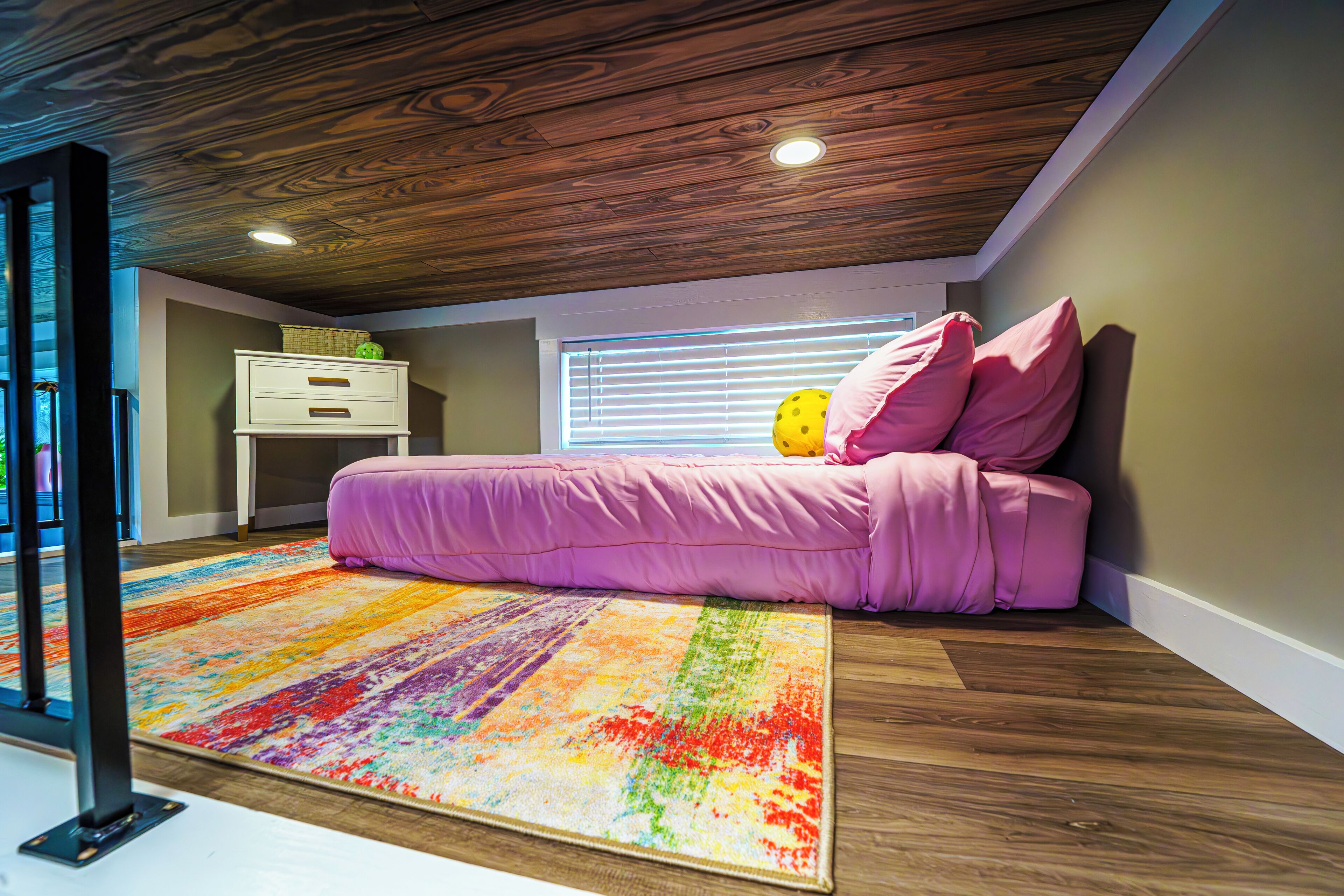 A bedroom with a wooden ceiling, a colorful rug, a bed with pink bedding, and a window with blinds.