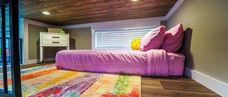 A bedroom with a wooden ceiling, a colorful rug, a bed with pink bedding, and a window with blinds.