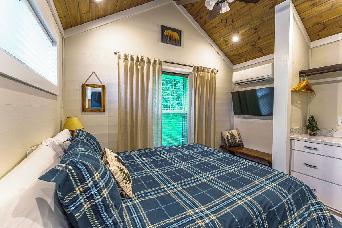 A bedroom with a bed, a window with curtains, a bedside table, a lamp, a bench, a small plant, and a wall-mounted mirror.