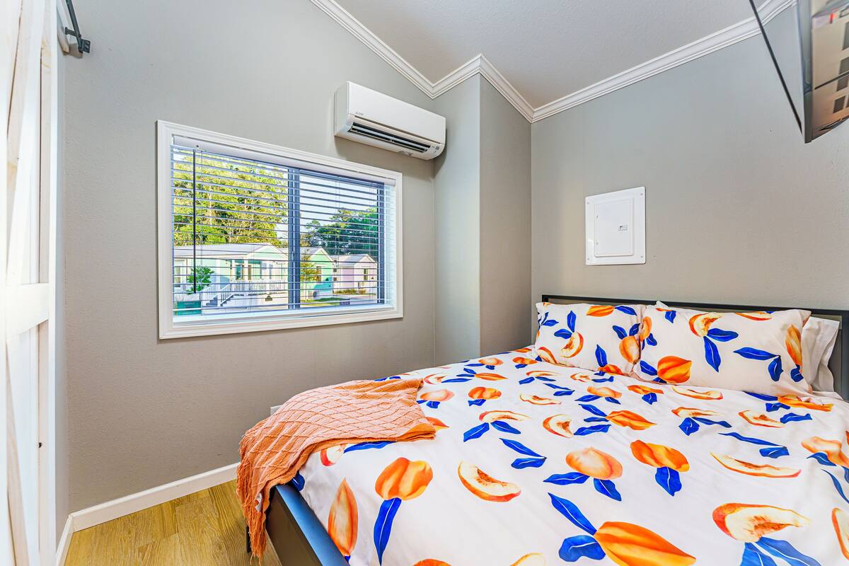 A bedroom with a bed, an air conditioning unit, a window with blinds, and a view of buildings outside.