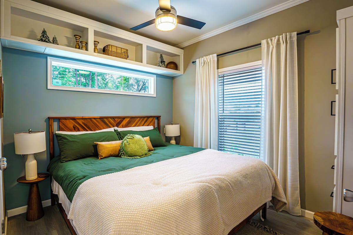 A bedroom with a bed, bedside lamps, a ceiling fan, a window with blinds, and a door.