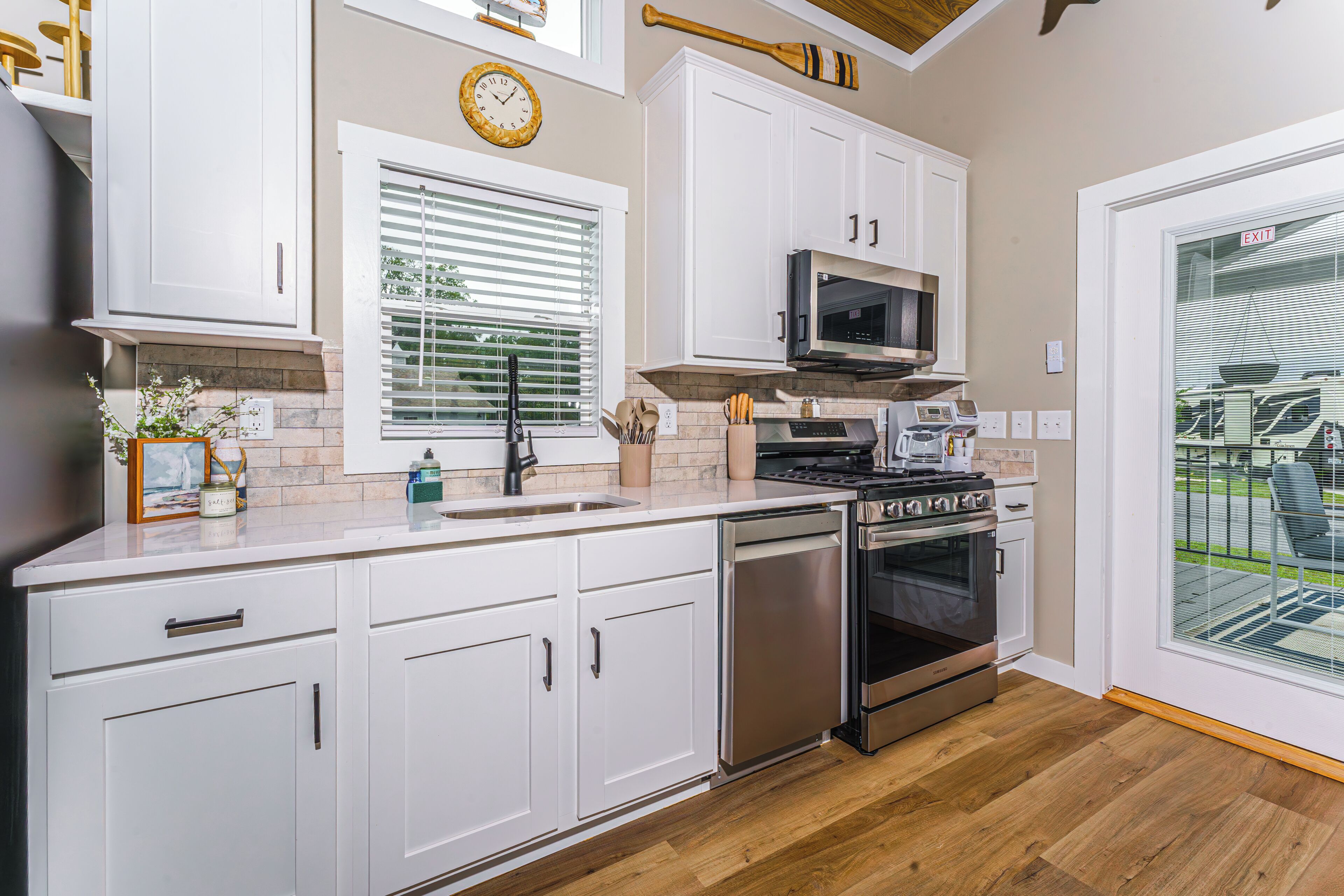 A modern kitchen with white cabinets, stainless steel appliances, a clock, and a view of the outdoors through a glass door.