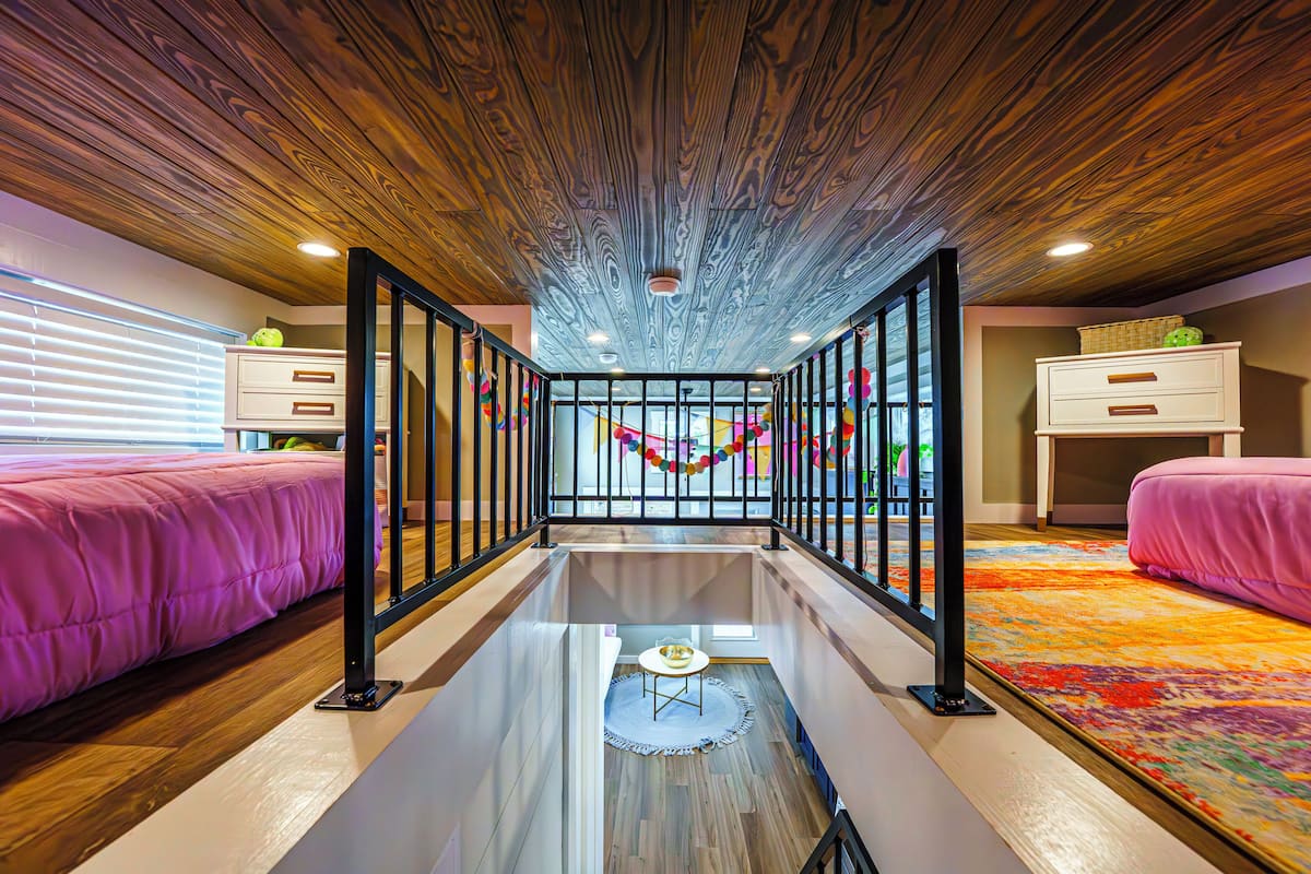 A room with a glass floor, two beds with pink bedding, a wooden ceiling, and a colorful carpet.