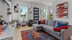 A modern living room with a grey sectional sofa, a colorful rug, and a large abstract painting. - Elevated Luxury 3BR • Mins to NRG • Med Center • Downtown • GRB (Houston)