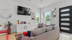 A modern living room with a grey sofa, a red chair, a TV mounted on the wall, and a black door with windows. - Elevated Luxury 3BR • Mins to NRG • Med Center • Downtown • GRB (Houston)