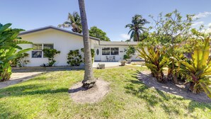 A single-story house with a white exterior, a front yard with grass and a palm tree, and a small patio area with outdoor furniture. - 1 Mi to Sand! Deerfield Beach Home w/ Patio (Deerfield Beach)