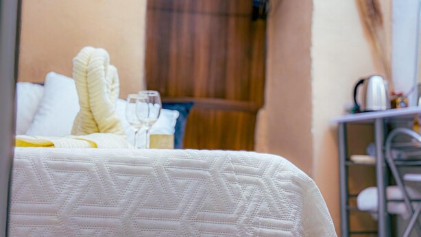 A neatly made bed with a patterned bedspread, a bedside table with a kettle and glasses, and a wooden door in the background. - roga bjeruslem (Jerusalem)