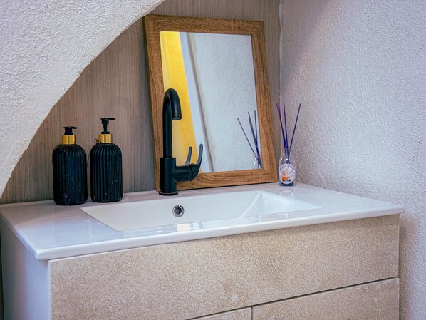 A bathroom vanity with a white sink, black soap dispensers, a black faucet, a wooden-framed mirror, and reed diffusers. - roga bjeruslem (Jerusalem)