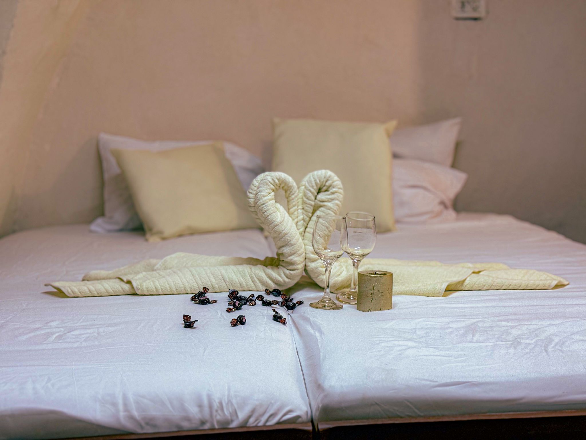 Cama bem arrumada com colcha branca, dois travesseiros, um arranjo decorativo de toalhas brancas, dois copos de vinho e uma vela.