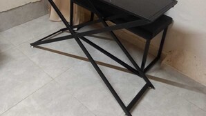 A corner dining table with a glass top and black metal frame, positioned against a wall with a power outlet. - roga bjeruslem (Jerusalem)