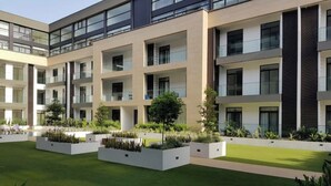Un immeuble moderne à plusieurs étages, doté de balcons, d’une cour avec pelouse et jardinières, et surplombant un ciel dégagé.
