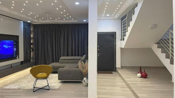 A modern living room with a flat-screen TV, grey sofa, yellow chair, and a staircase with a dark door.