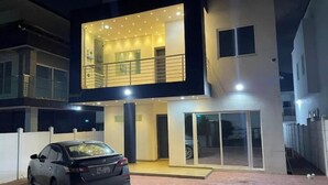 A modern two-story house with a balcony, a car parked in front, and a well-lit interior.