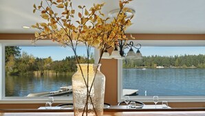 Une pièce dotée d’une grande fenêtre donnant sur un lac, un vase orné de branches et une table avec des verres.