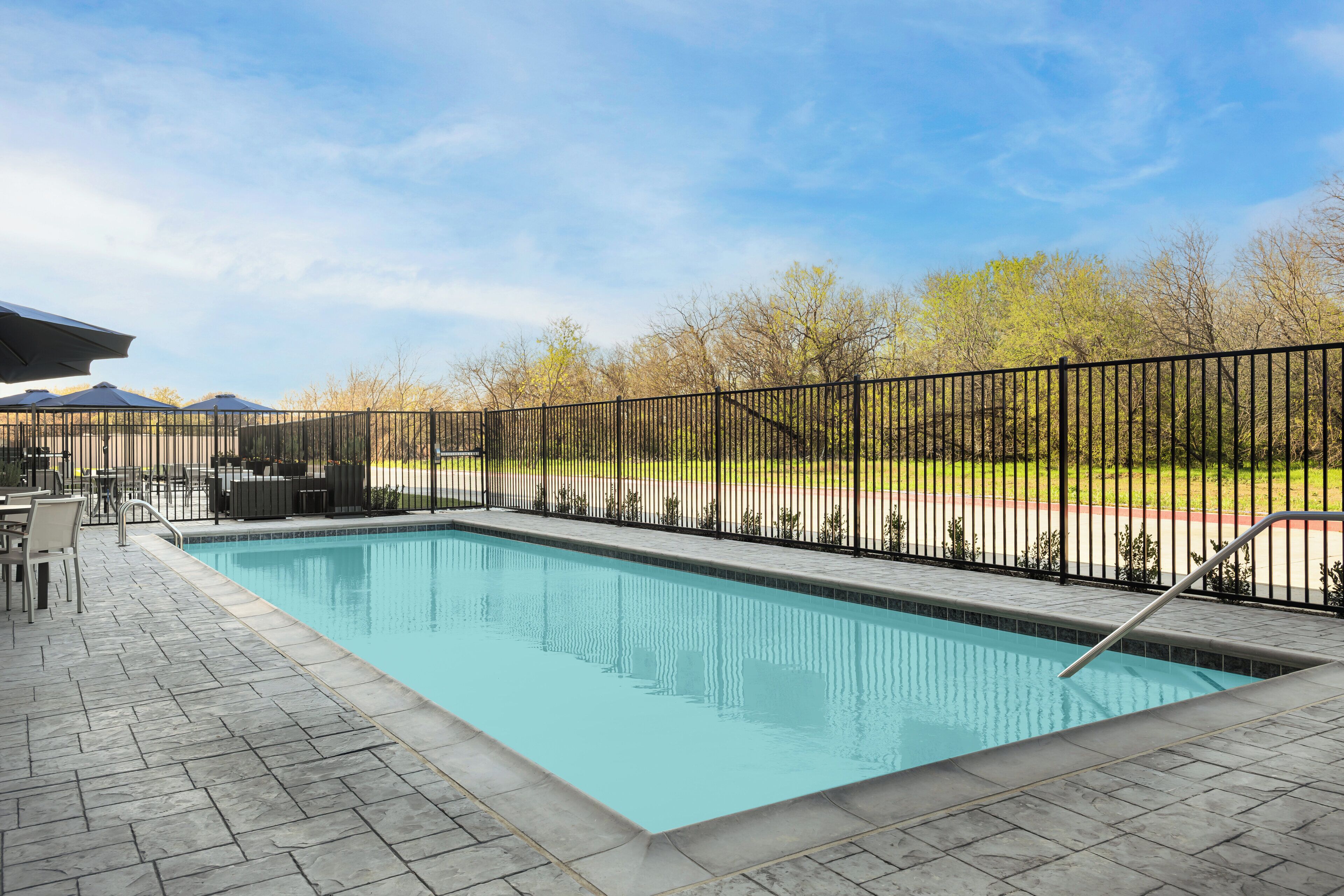 Una piscina rectangular con cerramiento de metal negro, solárium con baldosas y mobiliario exterior.