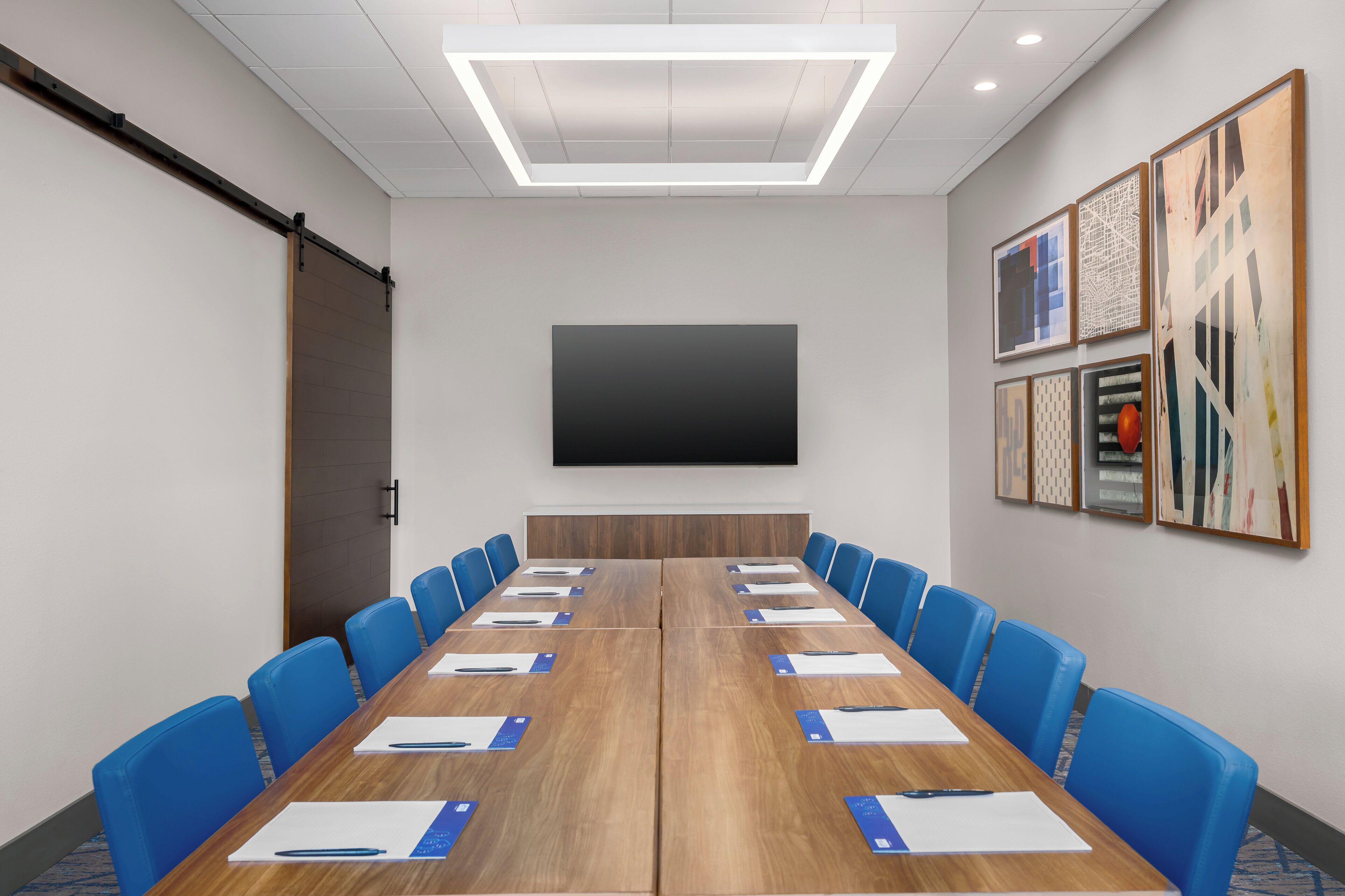A conference room with a long table, blue chairs, a large screen, and framed artwork on the walls.