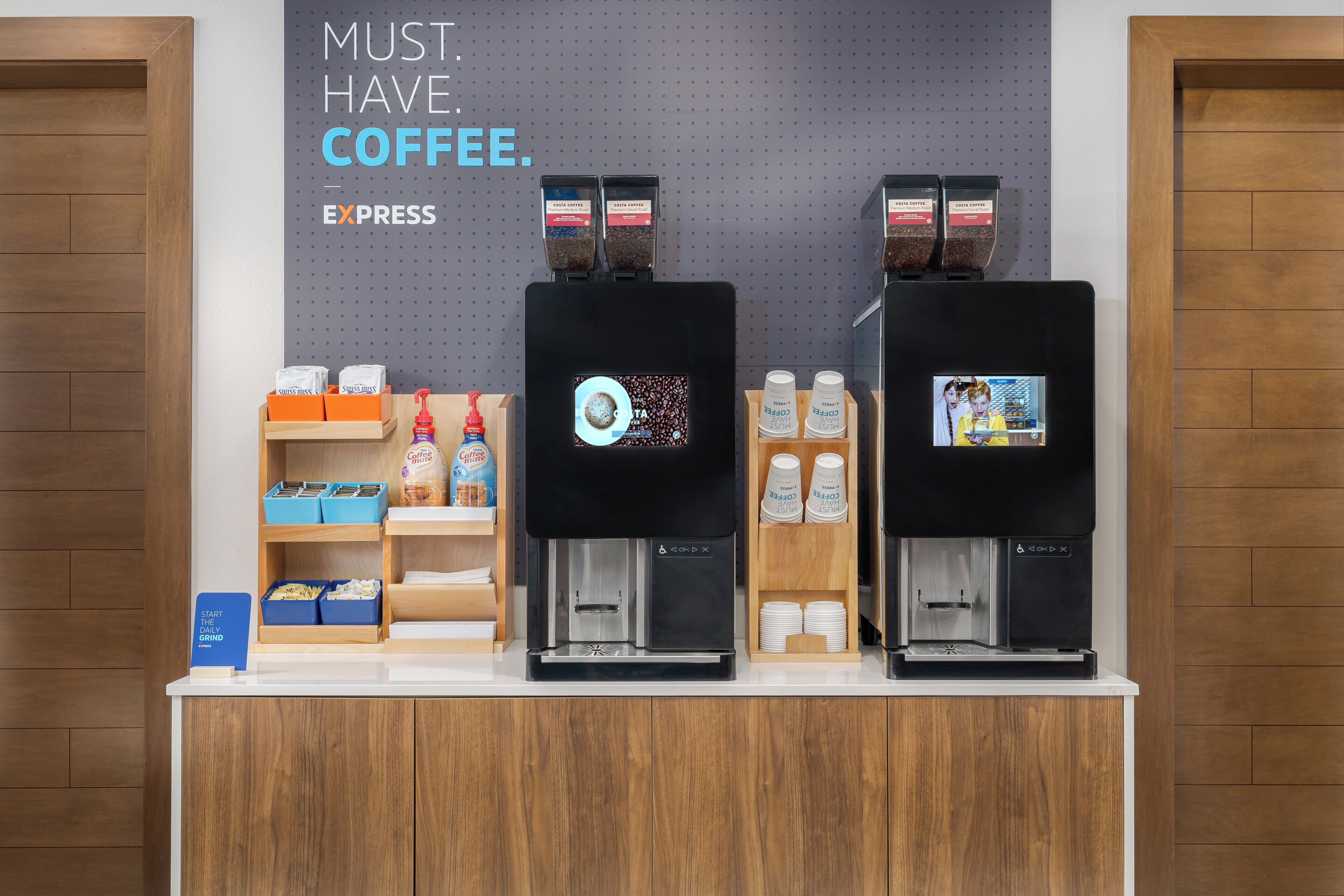 Un espace dédié au café, équipé de deux distributeurs de café, d’une pancarte indiquant « IL FAUT ABSOLUMENT DU CAFÉ » et proposant divers produits à base de café.