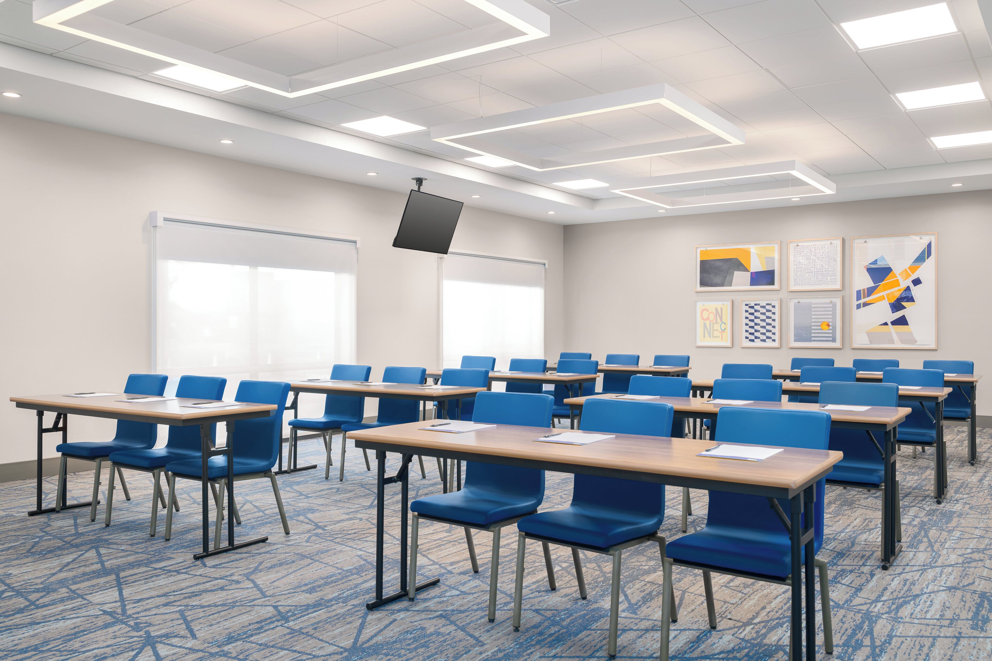 A conference room with rows of blue chairs and wooden tables, a whiteboard, and framed artwork on the walls.