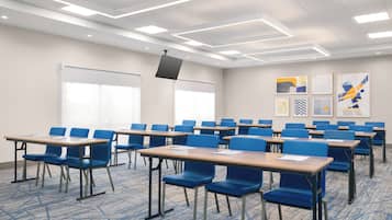 A conference room with rows of blue chairs and wooden tables, a whiteboard, and framed artwork on the walls.