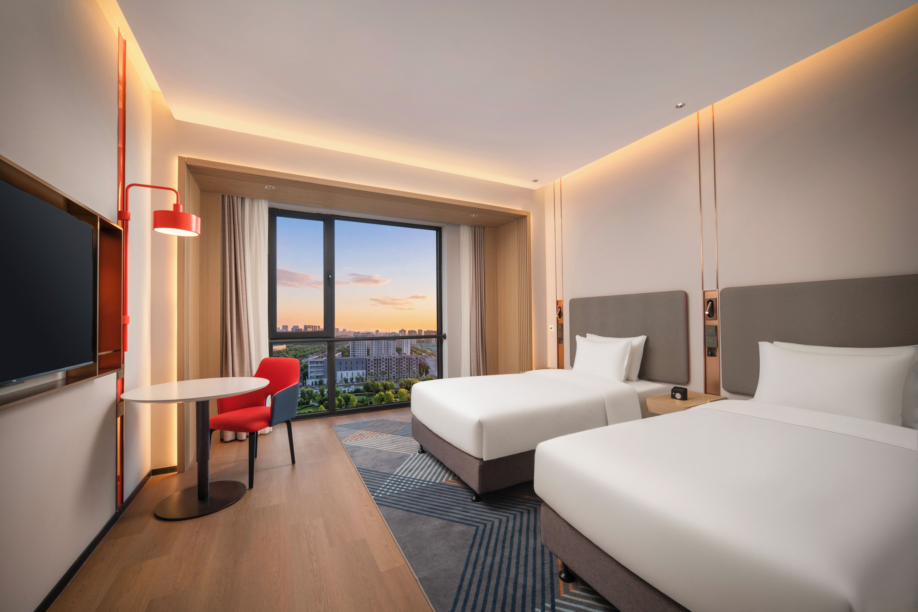 Habitación de hotel con dos camas, un escritorio, una silla, un televisor y un ventanal con vistas a la ciudad.